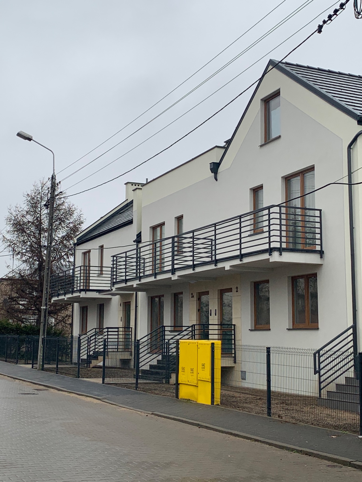 Nowo wybudowany szeregowiec z białymi elewacjami, czarnymi balustradami balkonowymi i schodami wejściowymi, sfotografowany w pochmurny dzień z perspektywy ulicy, z widoczną żółtą skrzynką gazową...