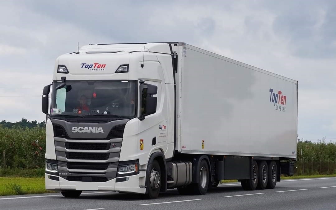 Biała ciężarówka Scania R450 z naczepą, logo TopTen Express na kabinie i naczepie, jadąca po asfalcie w pochmurny dzień.