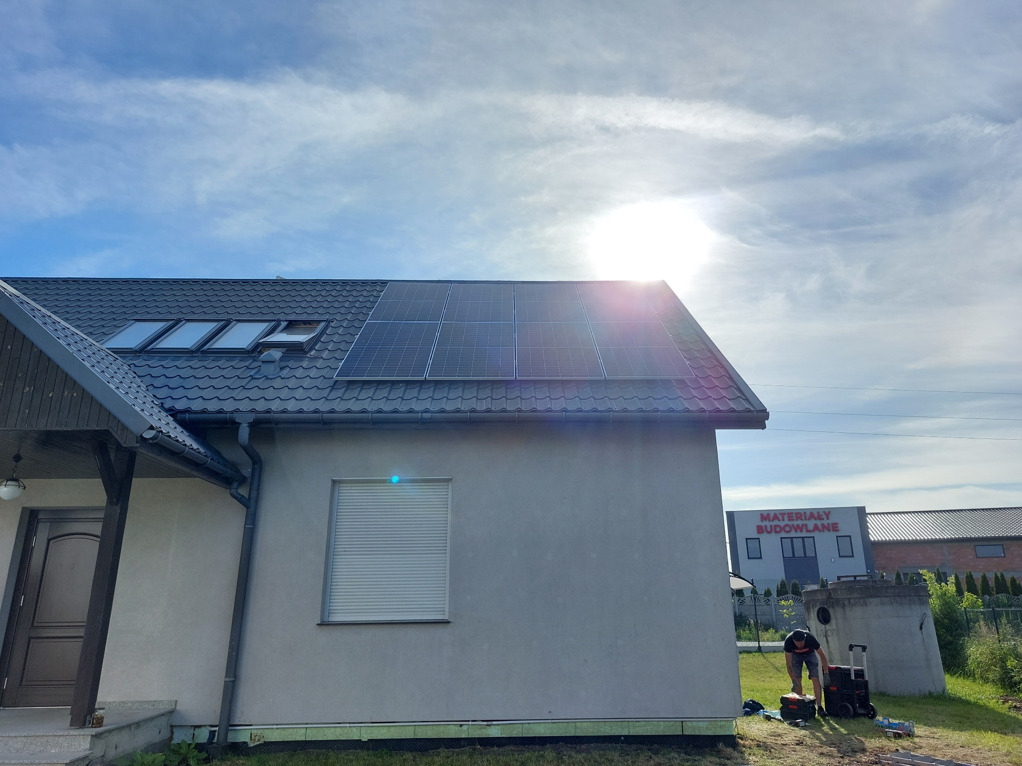Montaż paneli słonecznych na dachu domu jednorodzinnego we Wrocławiu, widoczny pracownik z narzędziami, w tle budynek z napisem 'Materiały Budowlane'.
