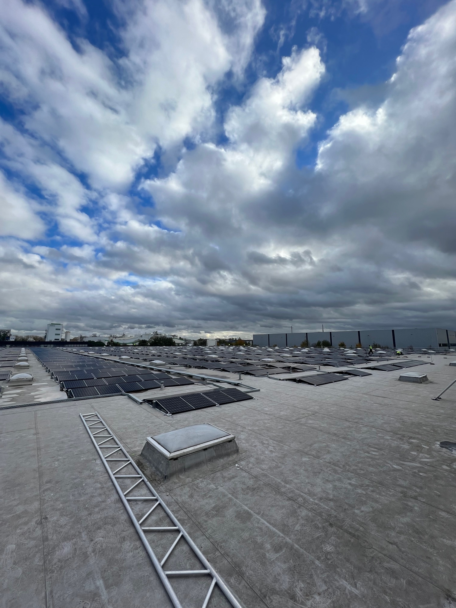 Widok z góry na dużą instalację paneli słonecznych na płaskim dachu budynku przemysłowego, z aluminiową drabiną leżącą obok świetlika dachowego i pochmurnym niebem w tle.