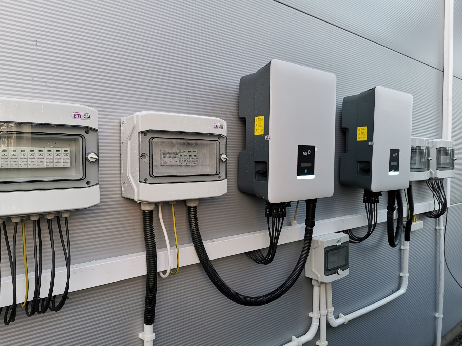 Dwa inwertery fotowoltaiczne marki FOX i dwie skrzynki rozdzielcze ETI zamontowane na zewnątrz budynku z szarej blachy trapezowej, widoczne przewody elektryczne.