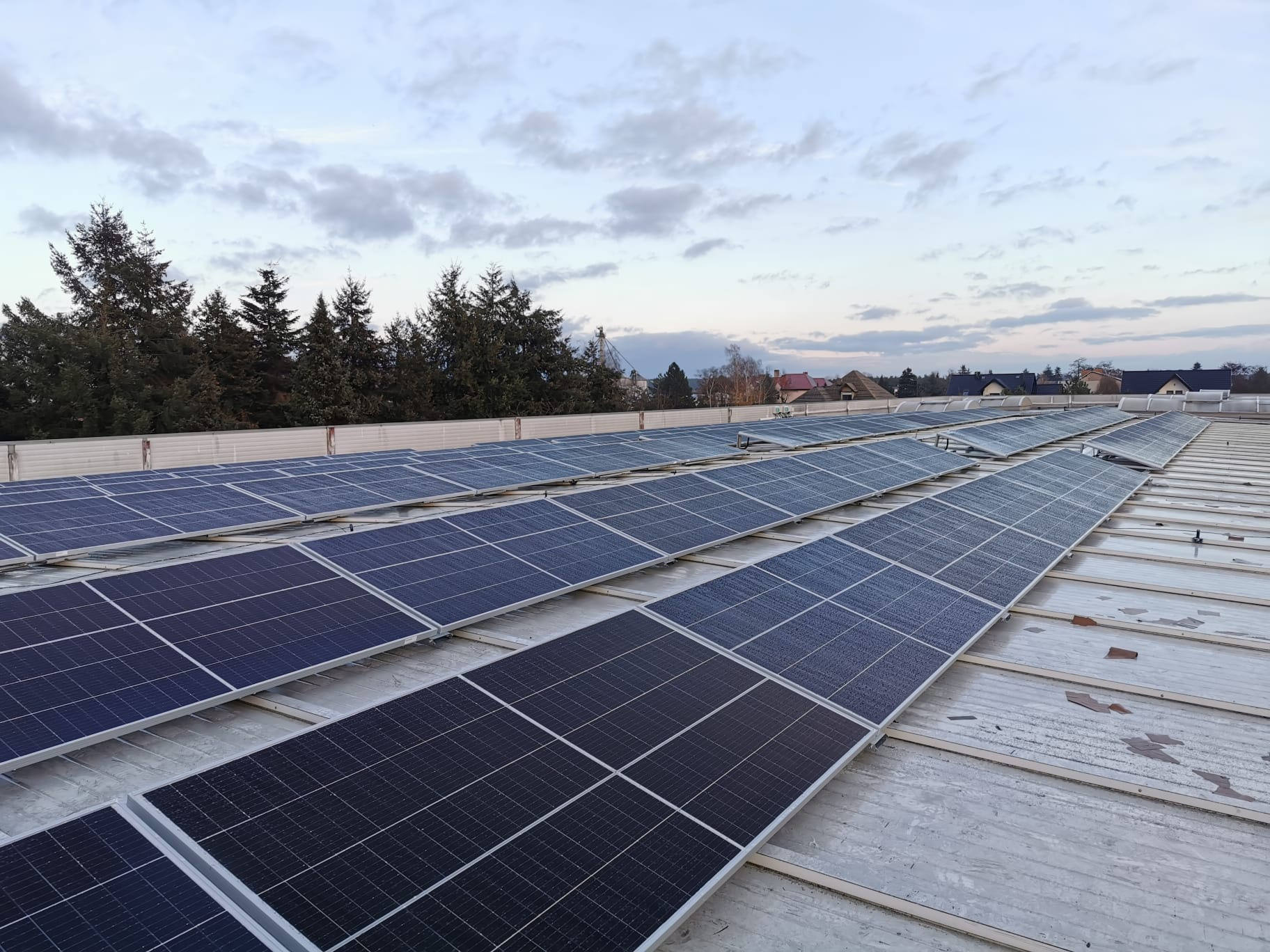 Rząd paneli słonecznych na dachu budynku, widok z góry na tle drzew i pochmurnego nieba.