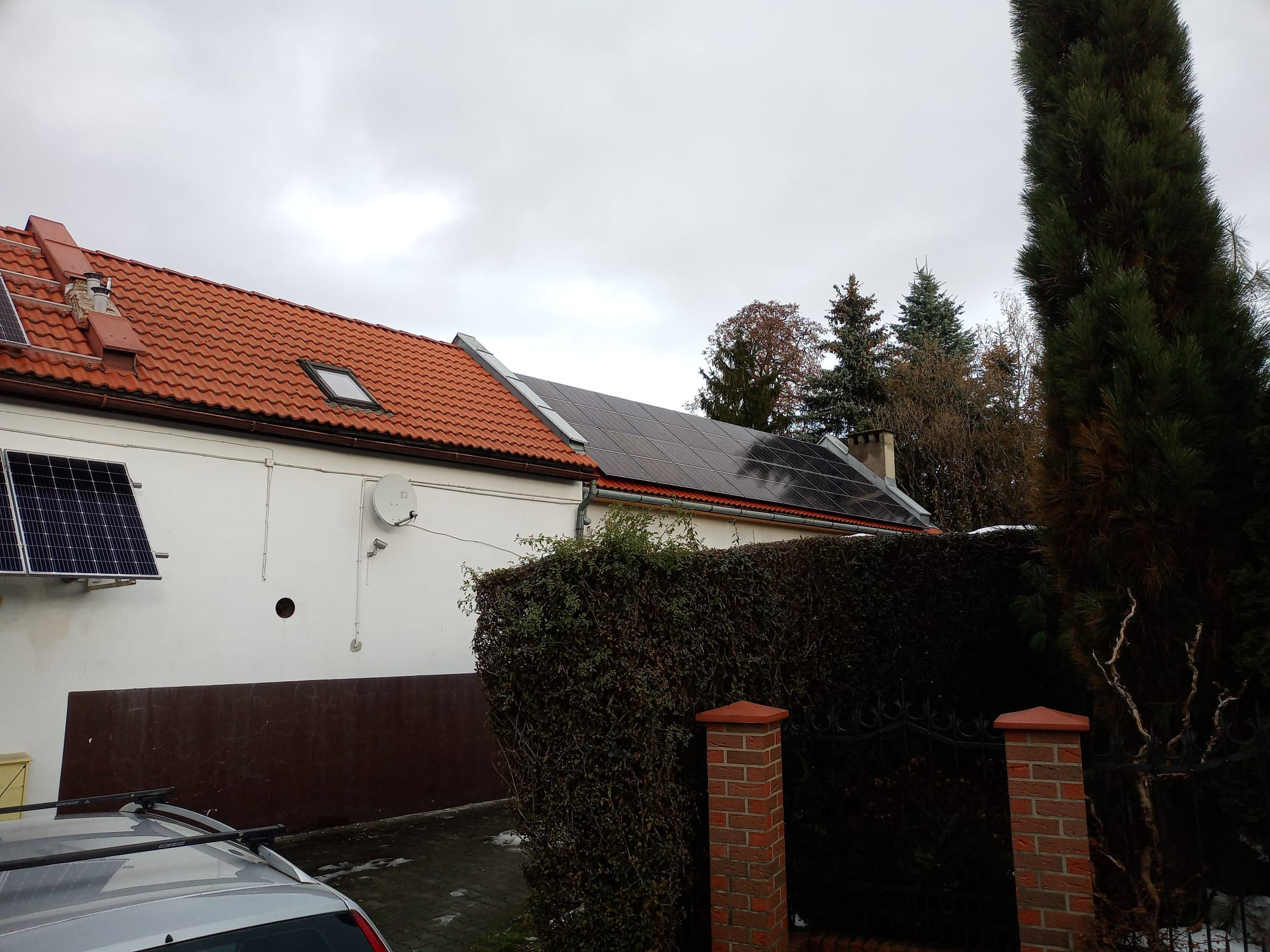 Dom z panelami słonecznymi na dachu, niektóre panele zamontowane na ścianie bocznej, widok z poziomu ulicy w pochmurny dzień.