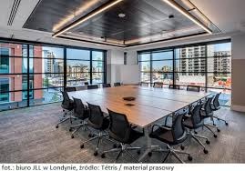 Nowoczesna sala konferencyjna z dużym drewnianym stołem, otoczona oknami z widokiem na miasto, oświetlona kwadratową lampą sufitową.