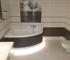 Nowoczesna łazienka z narożną wanną obudowaną mozaiką, oświetleniem LED pod wanną i białymi płytkami z ciemnobrązowymi akcentami.