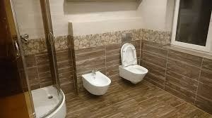 Nowoczesna łazienka z brązowymi płytkami imitującymi drewno, kabina prysznicowa z przezroczystego szkła, wisząca miska WC i bidet. Na ścianie dekoracyjny pas płytek z motywem kwiatowym. Okno...