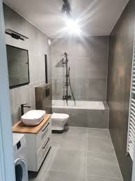 Nowoczesna łazienka w szarościach z czarnymi akcentami: bateria umywalkowa, rama lustra, deszczownica. Umywalka nablatowa na szafce z drewnianym blatem, obok pralka. Widoczna wanna z parawanem...