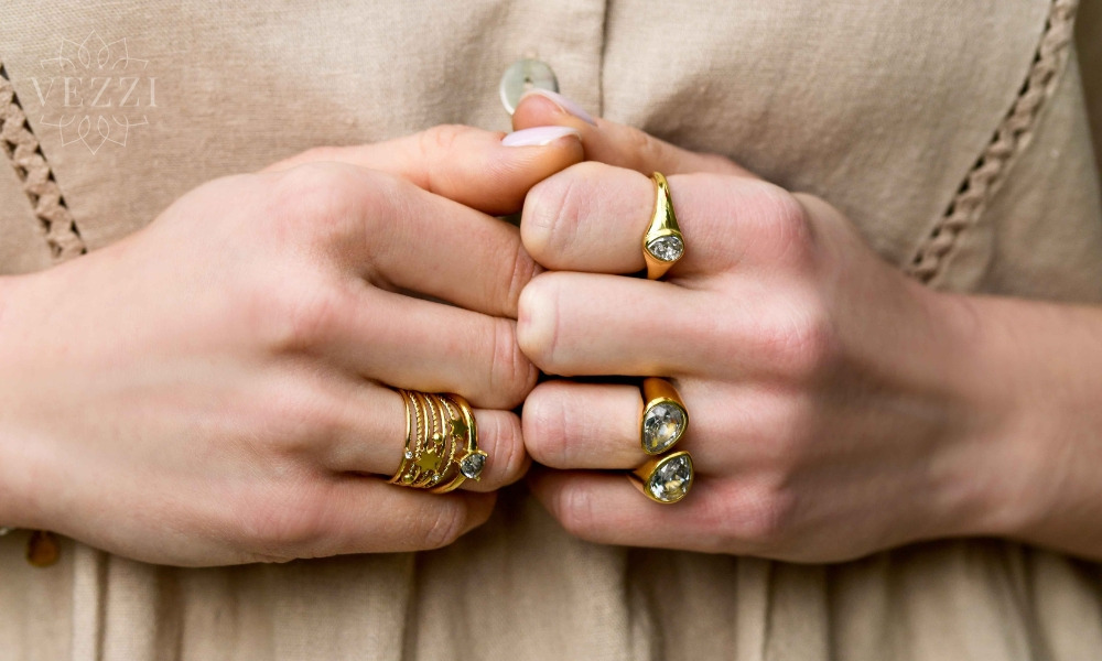 Dłonie kobiety z delikatnym manicure prezentującej złote pierścionki z jasnymi kamieniami na tle beżowej bluzki.