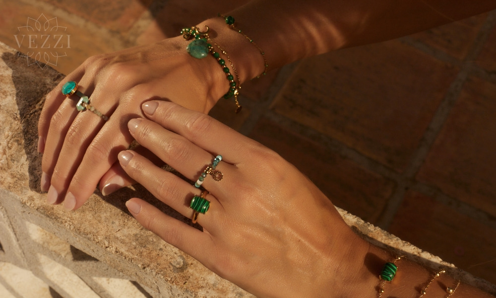 Dłonie modelki prezentującej złotą biżuterię z zielonymi kamieniami: pierścionki z malachitem i turkusem oraz bransoletki z zawieszkami, na tle kamiennej powierzchni.