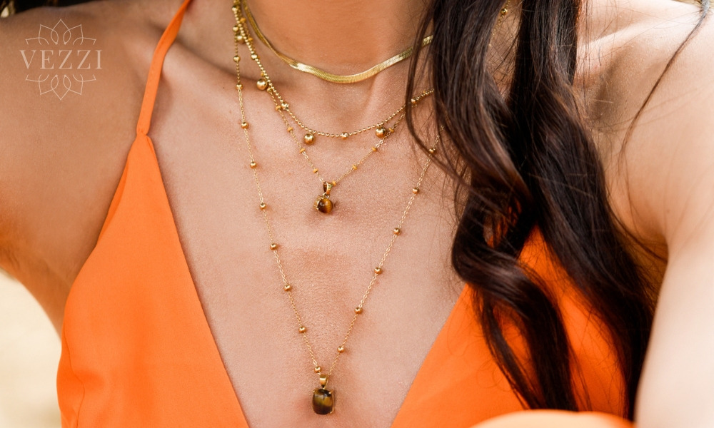 Kobieta w pomarańczowej sukience prezentuje zestaw złotych naszyjników o różnej długości i fakturze, w tym jeden z zawieszką z kamieniem tygrysie oko.