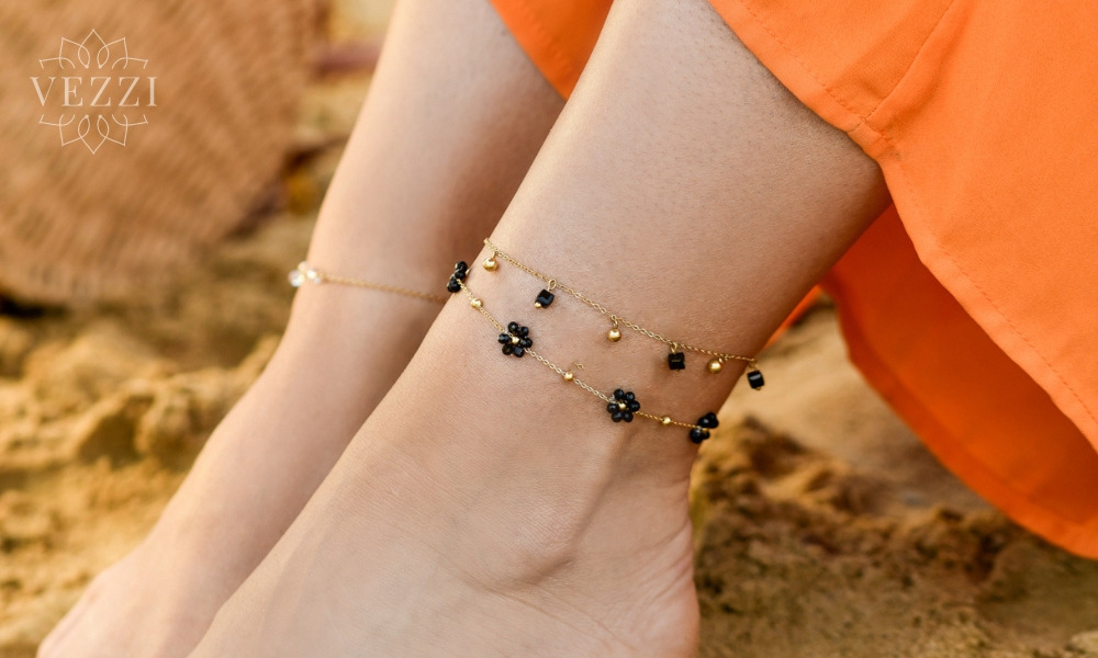 Dwie złote bransoletki na kostkę: jedna z czarnymi kwiatkami i złotymi koralikami, druga z perłowymi koralikami, na tle piasku i pomarańczowej sukienki.