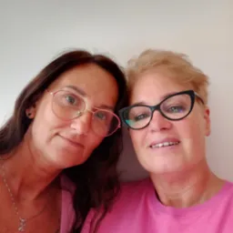 Portret dwóch uśmiechniętych kobiet w różowych koszulkach i okularach, przytulonych do siebie, z bliska na tle jasnej ściany.
