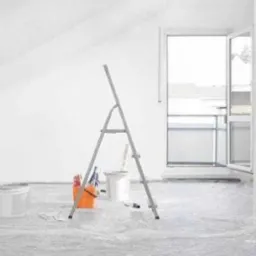 Pokój po malowaniu na biało, zabezpieczona folią podłoga, drabina, wiadro z narzędziami malarskimi i otwarte drzwi balkonowe.