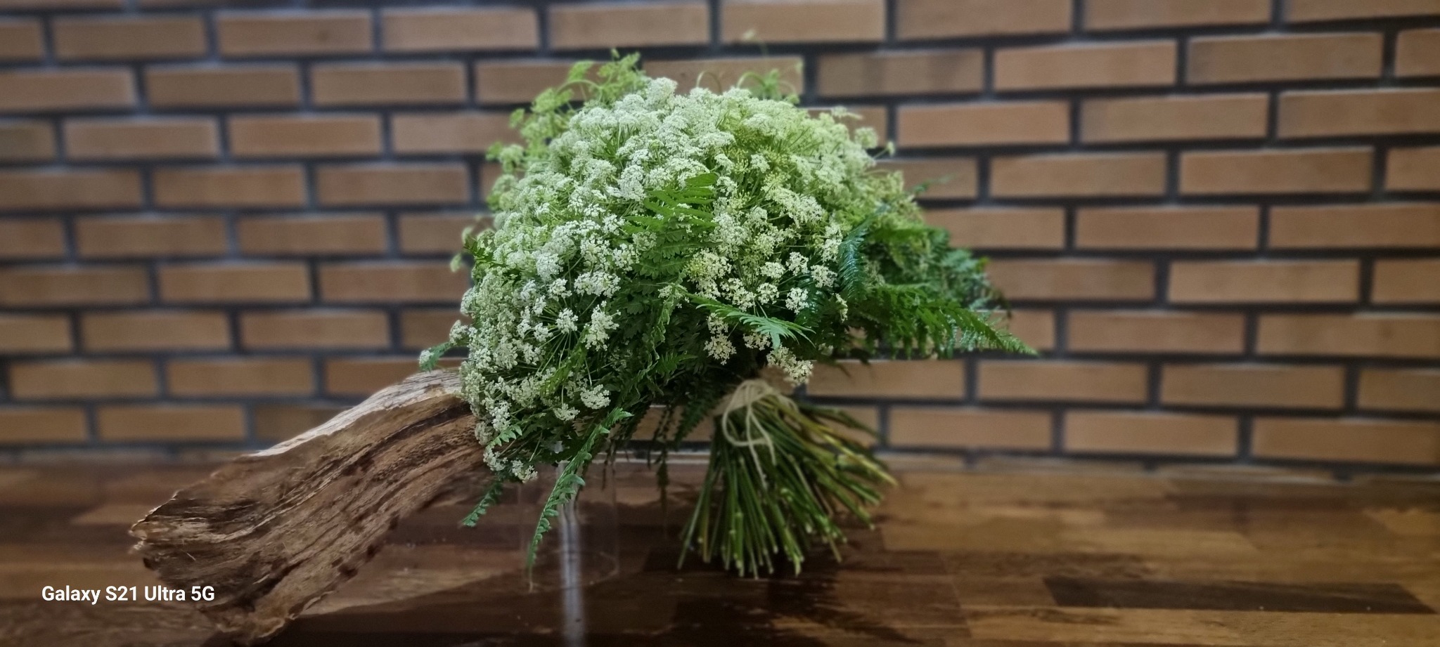 Dekoracyjny bukiet z białych kwiatów i paproci, związany sznurkiem, leżący na kawałku drewna na tle ściany z cegieł.