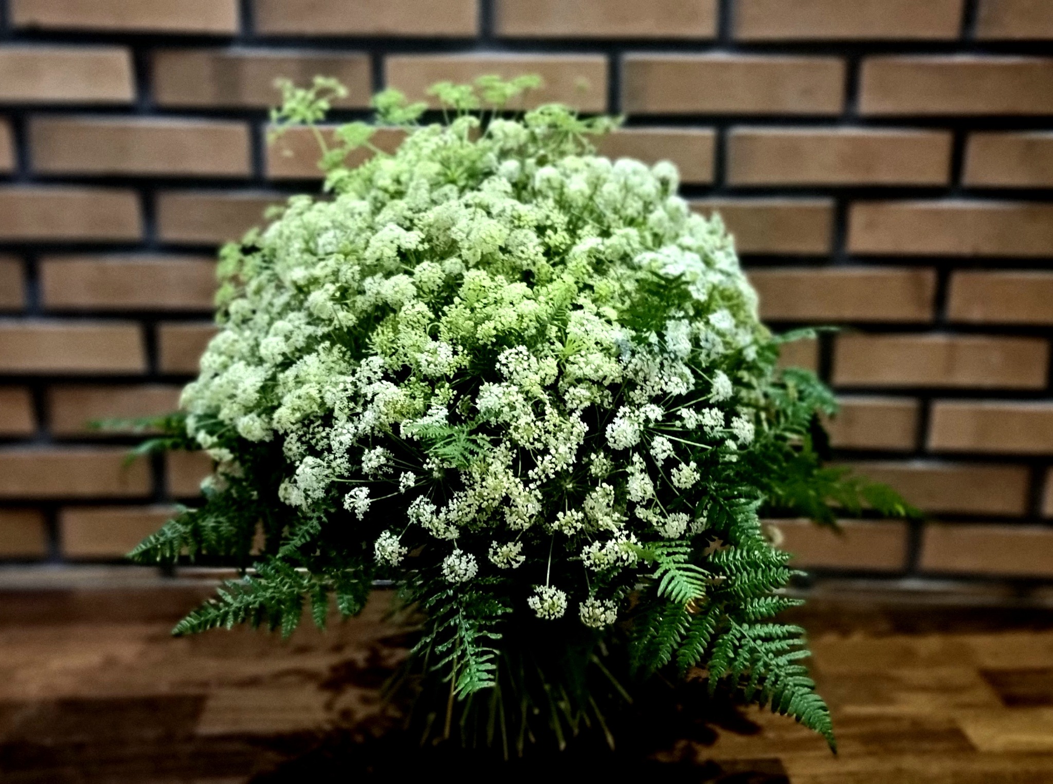 Gęsty bukiet z białych drobnych kwiatów i zielonych paproci na tle ceglanej ściany i drewnianego blatu.