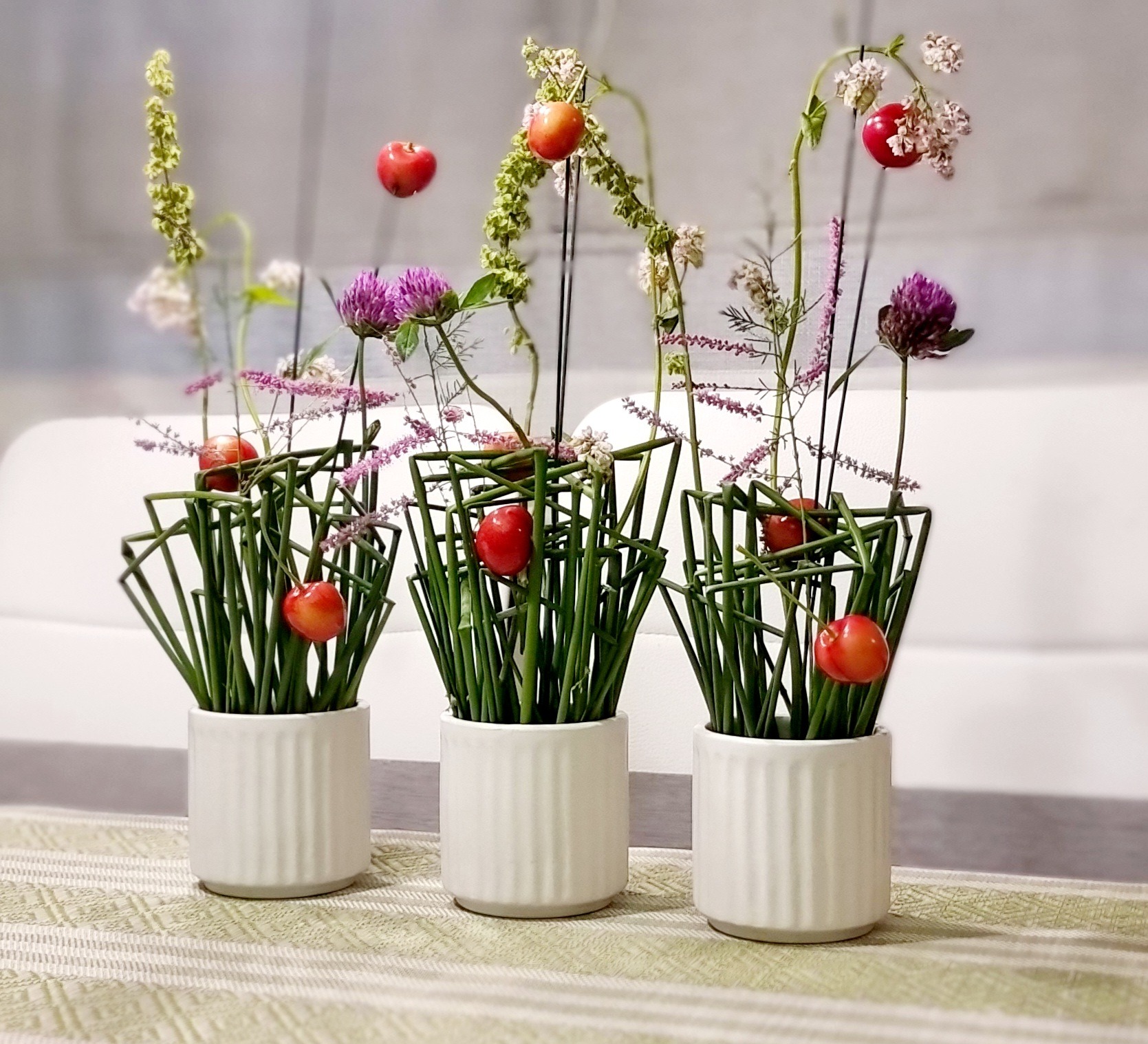 Trzy białe, prążkowane doniczki z nowoczesnymi kompozycjami kwiatowymi, w których dominują zielone źdźbła, fioletowe kwiaty i czerwone owoce, ustawione na wzorzystym obrusie.