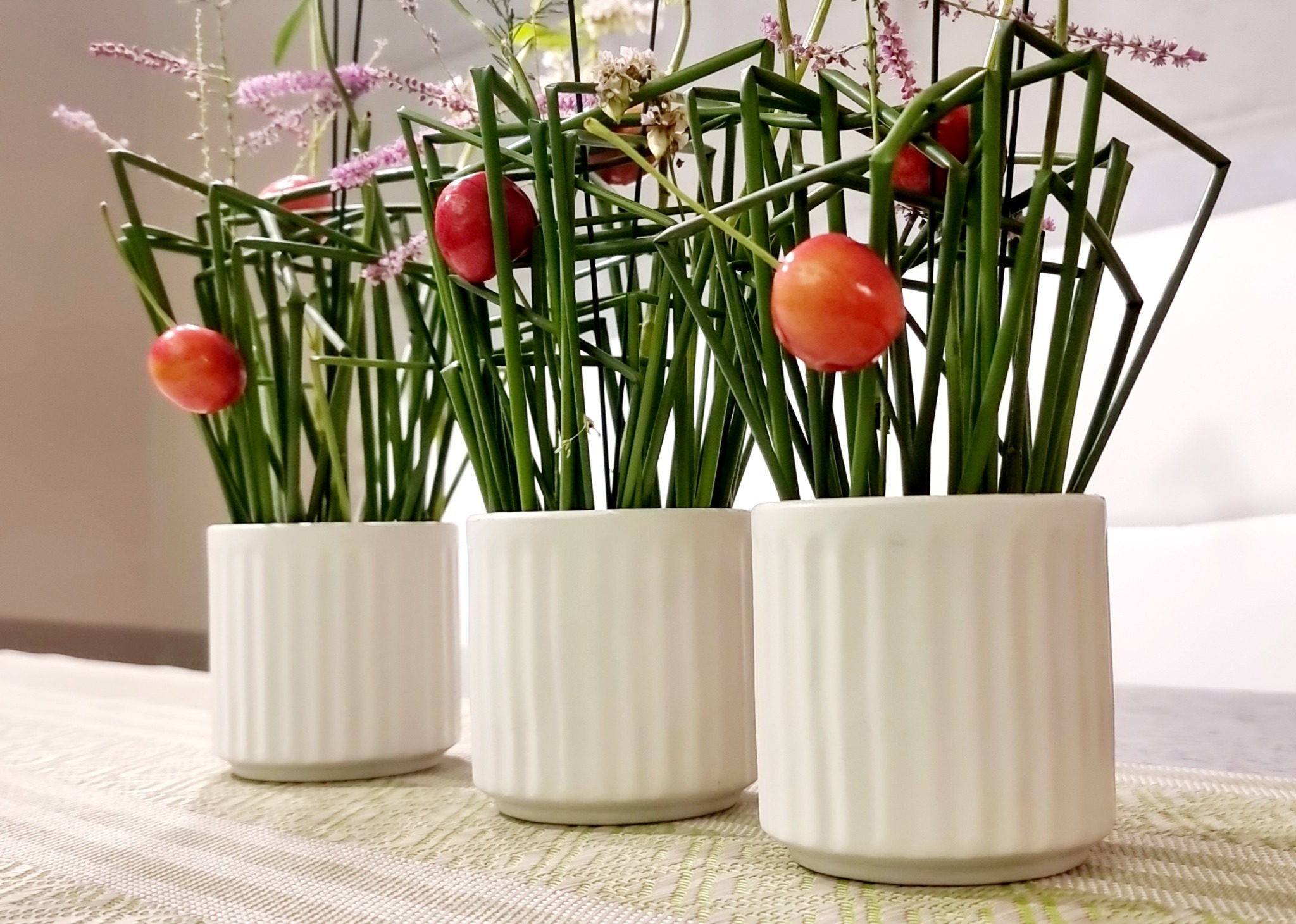 Trzy białe, żłobkowane doniczki z minimalistyczną kompozycją z zielonych łodyg, geometrycznych czarnych ramek i czerwonych owoców, ustawione na beżowym obrusie w paski.