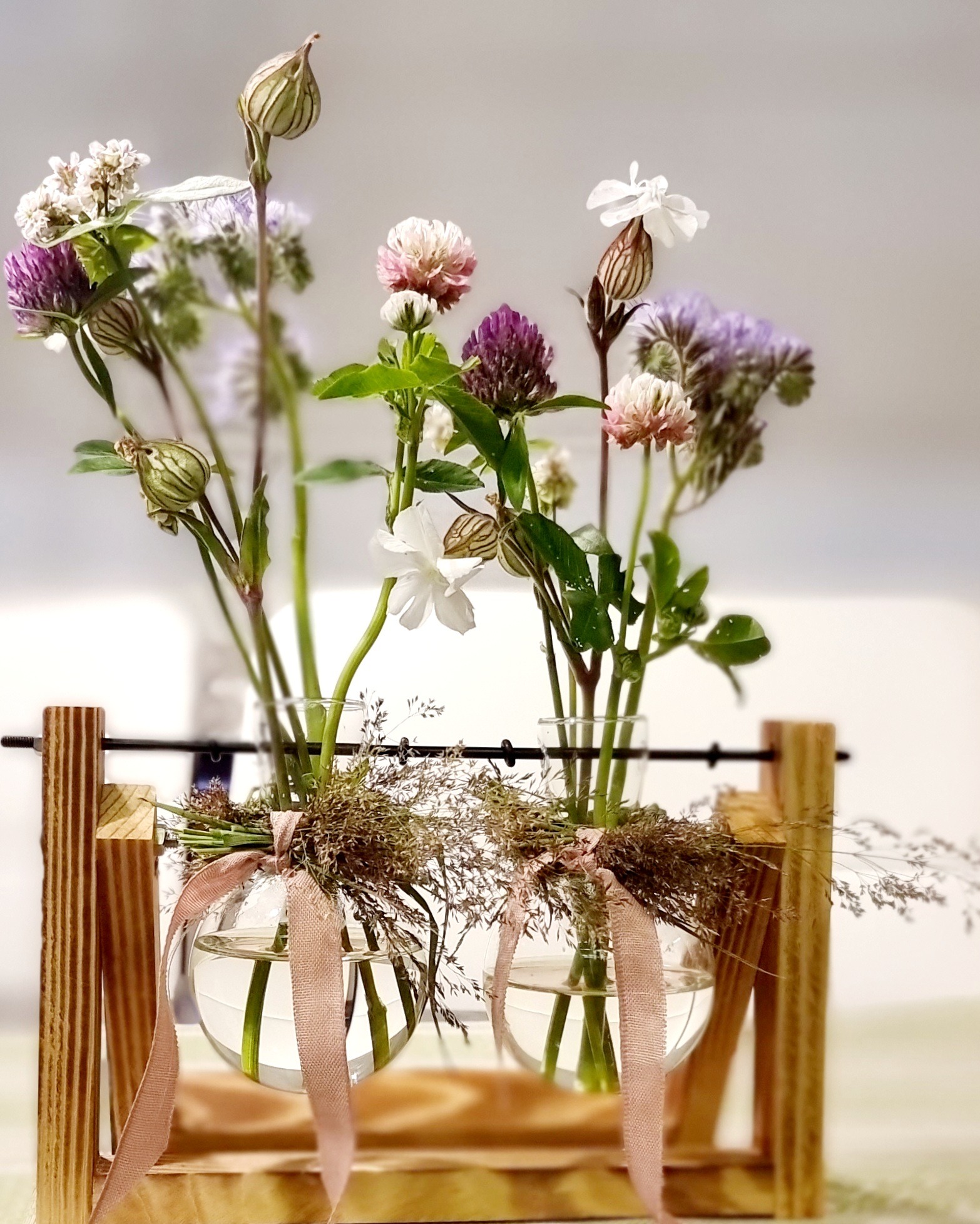 Dwa szklane wazony kuliste zawieszone w drewnianej konstrukcji, wypełnione polnymi kwiatami i przewiązane różową wstążką.