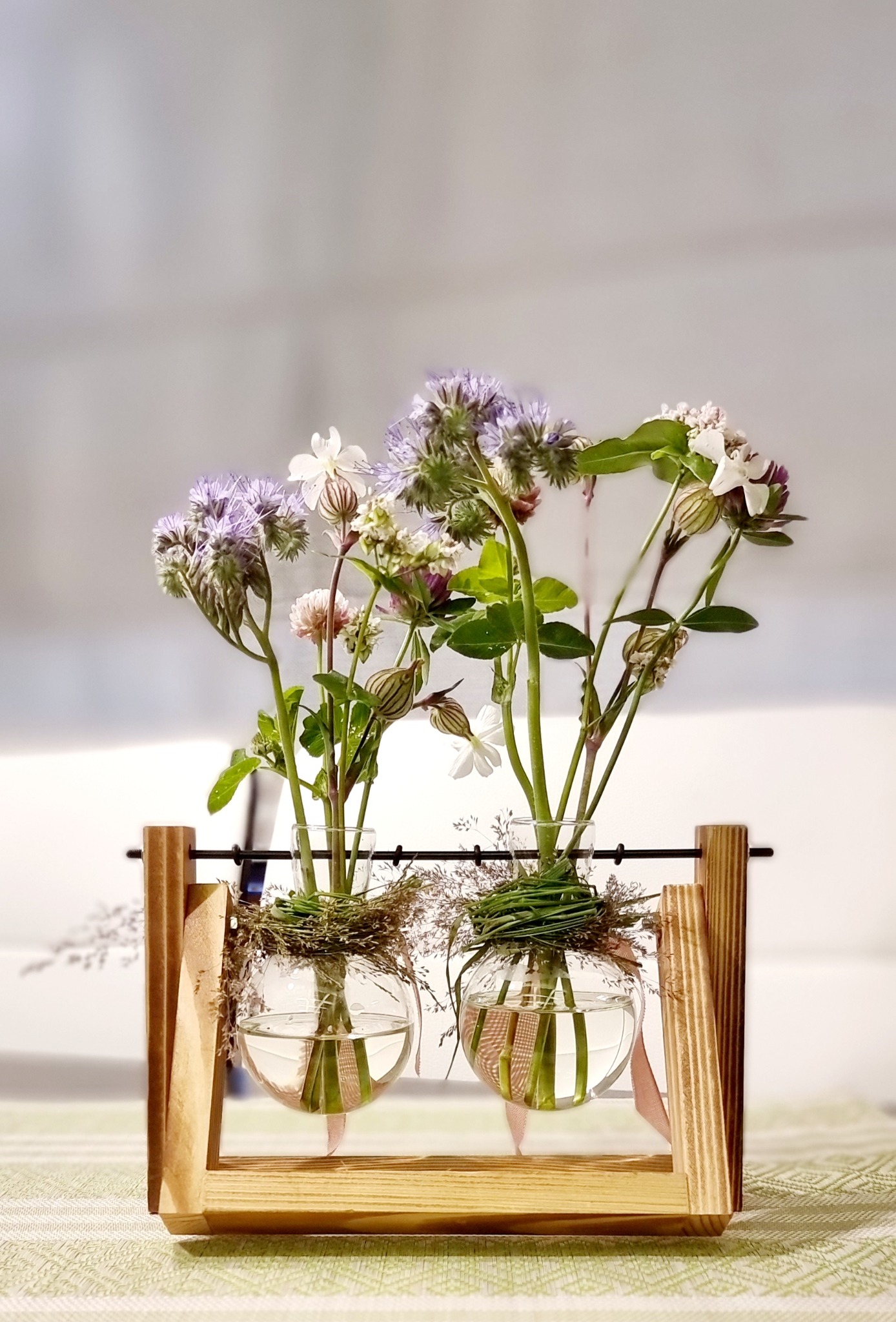 Dwa okrągłe wazoniki z fioletowymi i białymi kwiatami, umieszczone w drewnianej ramce, ozdobione trawą i wstążką, na stole w delikatne wzory.