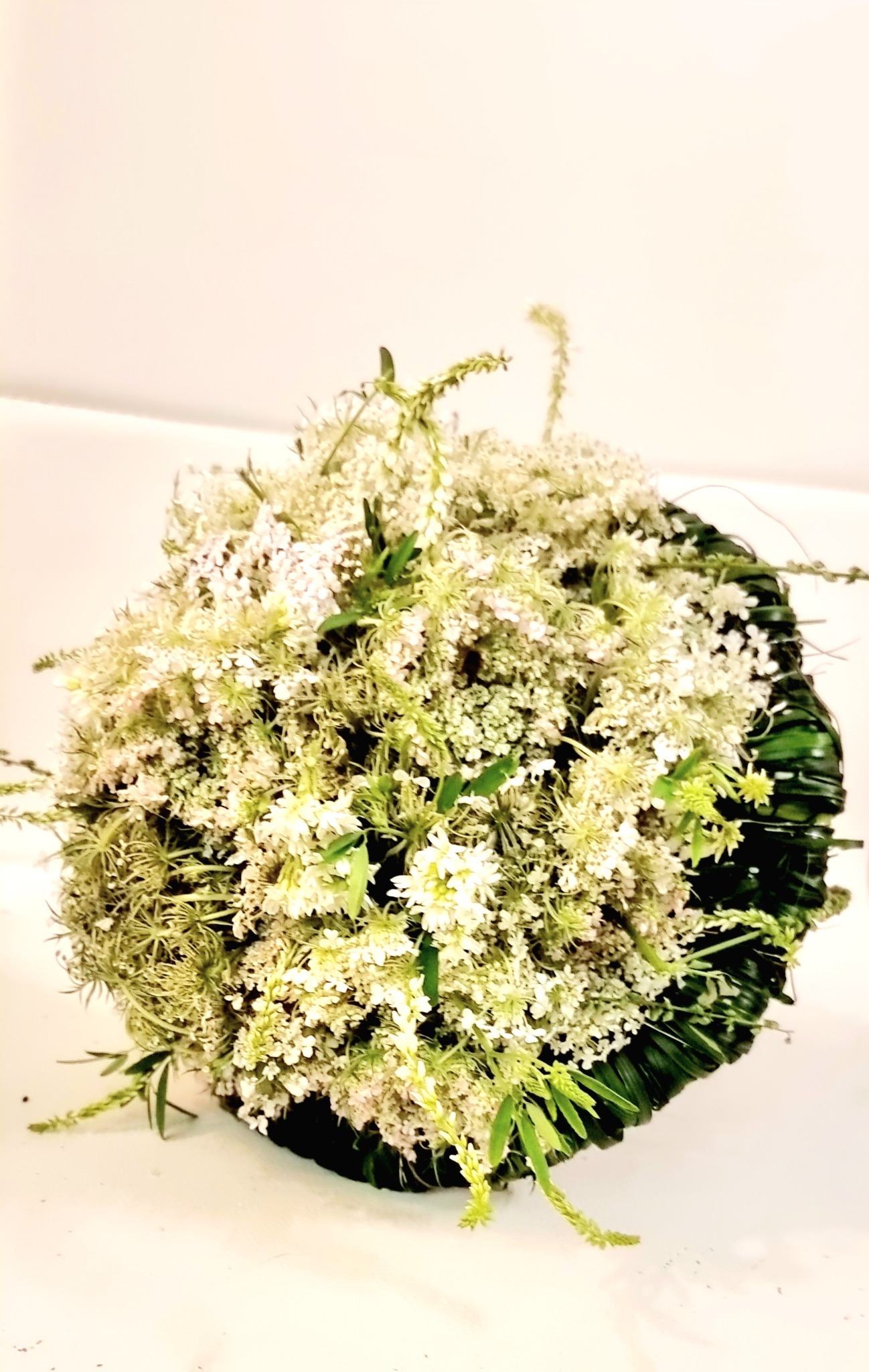 Gęsty bukiet z drobnych białych kwiatów i zielonych liści, ułożony w kulistej kompozycji z ciemnozieloną bazą.
