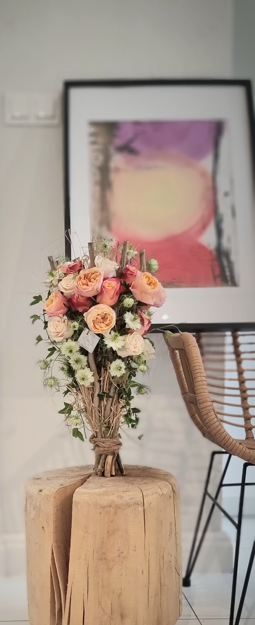 Bukiet z róż w odcieniach brzoskwini i bieli, z dodatkiem zielonych akcentów, umieszczony na drewnianym postumencie w jasnym wnętrzu z abstrakcyjnym obrazem w tle.