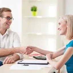 Uśmiechnięty mężczyzna w okularach w białej koszuli ściska dłoń uśmiechniętej blondynce w niebieskiej sukience nad biurkiem z kalkulatorem i dokumentami.