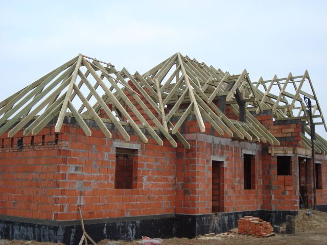 Niedokończony, jednopiętrowy budynek z czerwonej cegły z drewnianą konstrukcją dachu, widok z zewnątrz w pochmurny dzień.