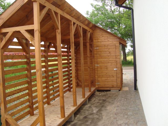 Drewniana konstrukcja z zadaszeniem, prawdopodobnie wiata lub część budynku gospodarczego, z widoczną konstrukcją szkieletową i poziomymi deskami tworzącymi ścianę, przylegająca do białej ściany...