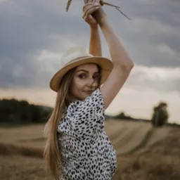 Portret kobiety w sukience w kropki i kapeluszu, trzymającej kłosy zboża na tle pola i pochmurnego nieba.