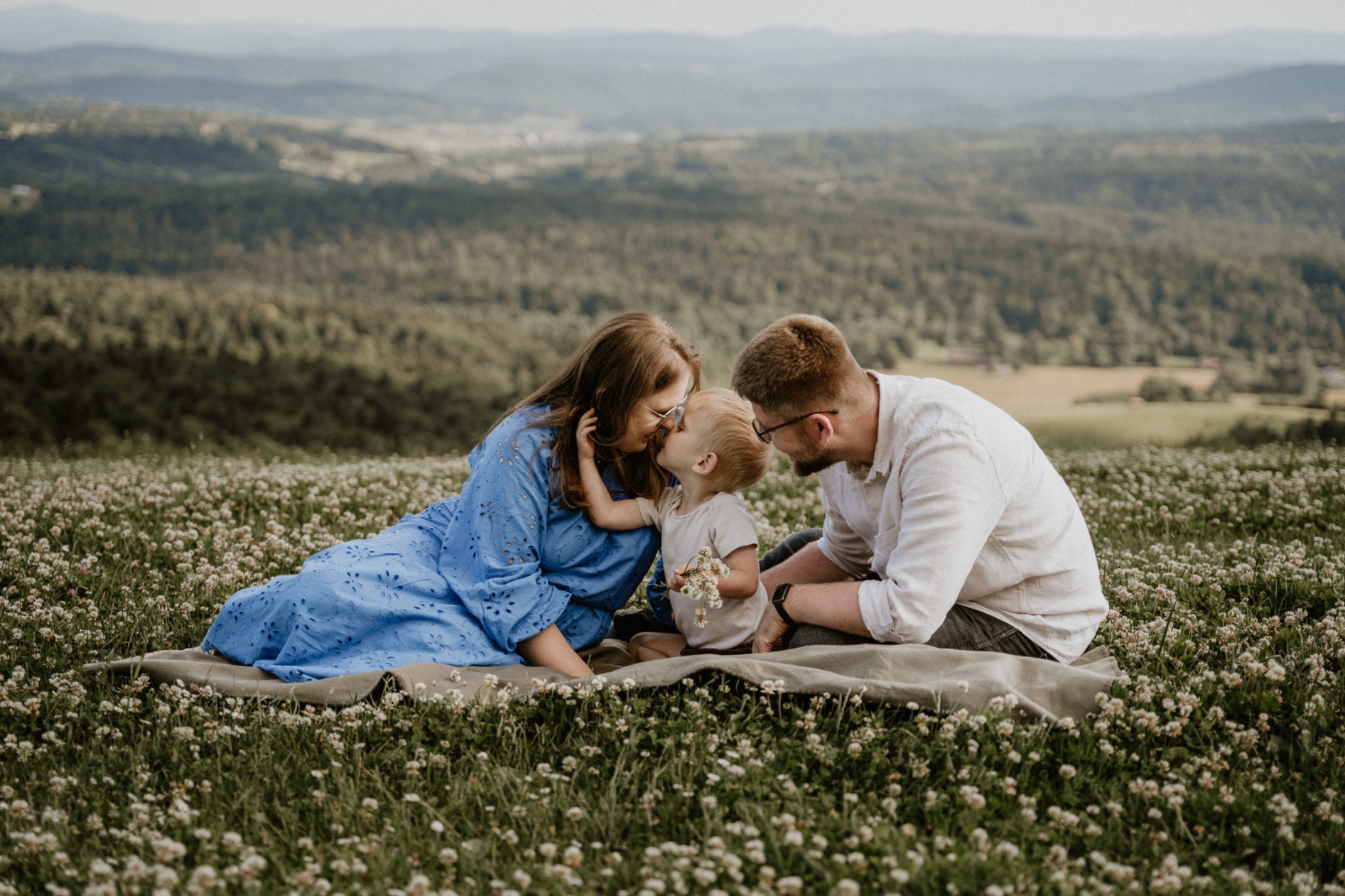 Rodzina (matka w niebieskiej sukience, ojciec w białej koszuli i syn) siedzi na kocu w kwitnącej łące z widokiem na zalesione wzgórza, dziecko trzyma bukiecik polnych kwiatów.