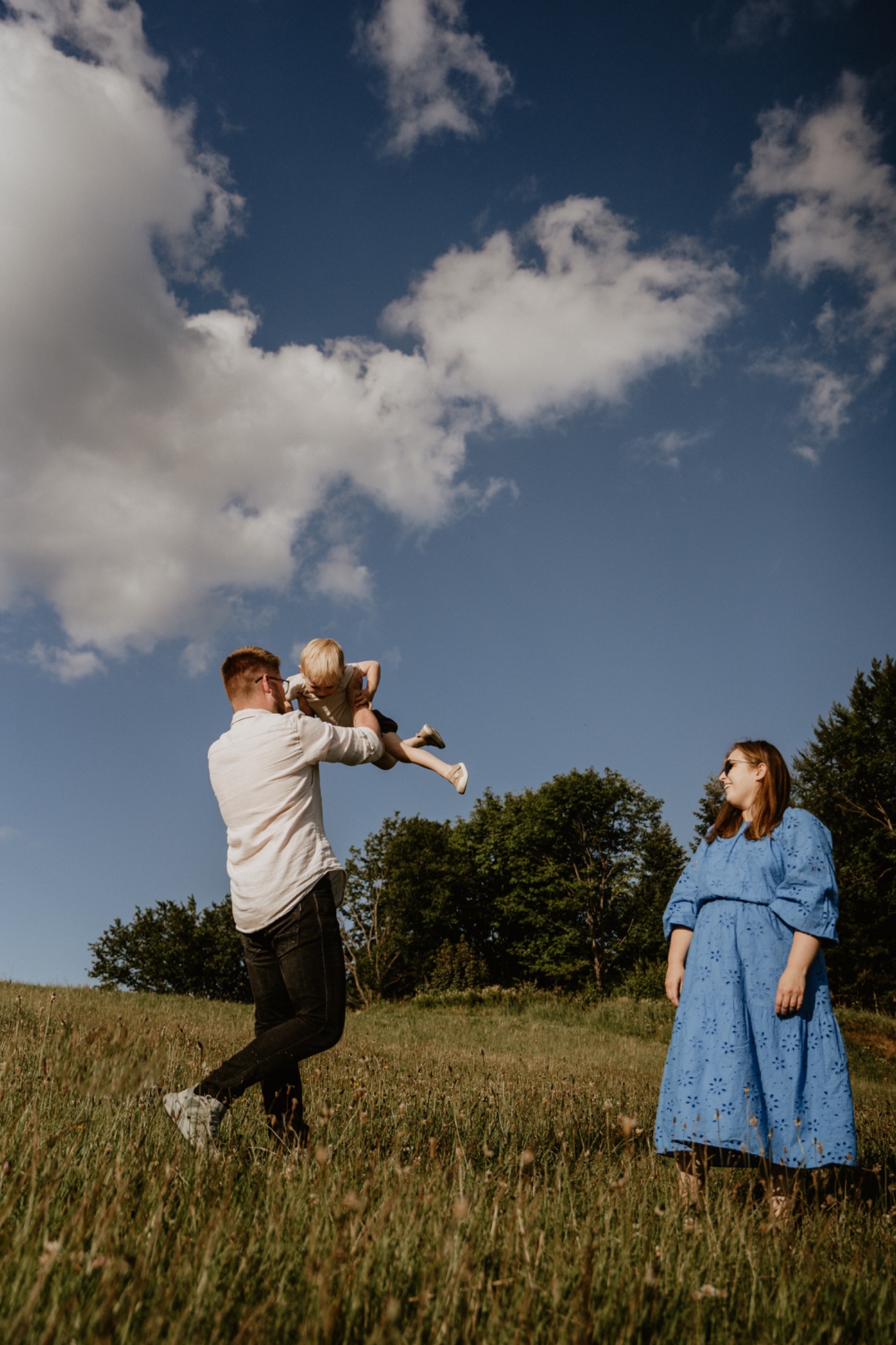 Ojciec podrzuca syna w górę na łące, matka w niebieskiej sukience obserwuje w słoneczny dzień.