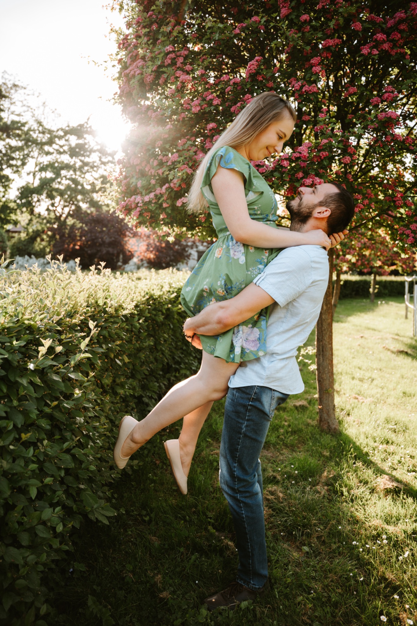 Uśmiechnięta para w objęciach na tle kwitnącego drzewa, mężczyzna trzyma kobietę w górze, delikatne światło słoneczne.
