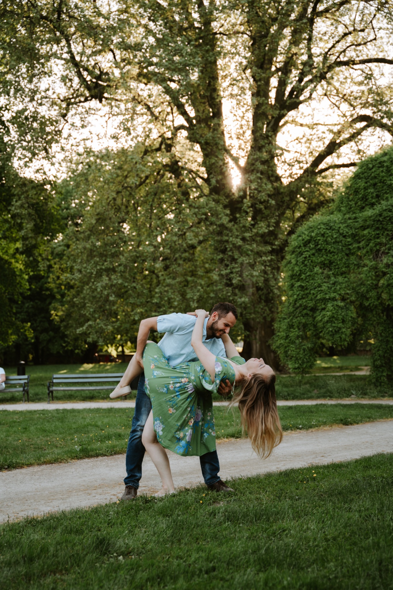 Uśmiechnięta para w parku: mężczyzna podtrzymuje kobietę w kwiecistej sukience, pochyloną do tyłu, z promieniami słońca przebijającymi przez korony drzew w tle.