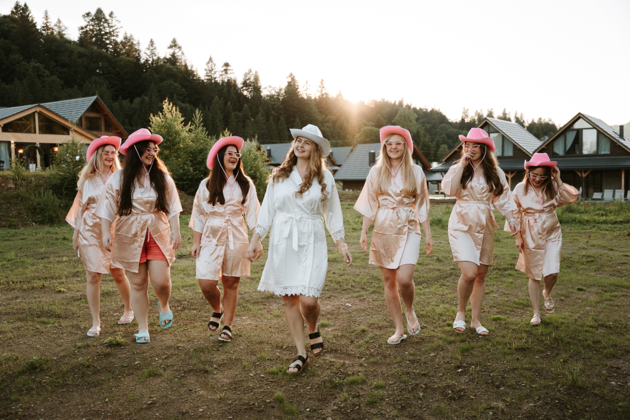 Grupa kobiet w szlafrokach i kowbojskich kapeluszach spaceruje po trawiastym terenie przed domem w górach, panna młoda w białym stroju i kapeluszu.
