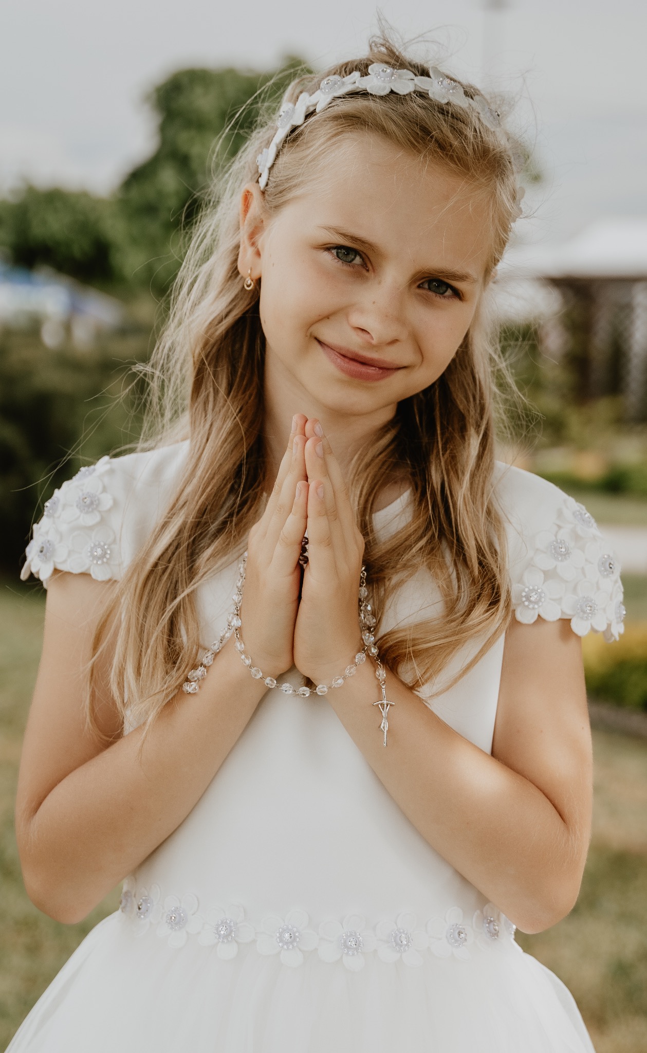 Dziewczynka w białej sukience komunijnej z różańcem w dłoniach, uśmiechnięta, z wiankiem na głowie, na tle zieleni.