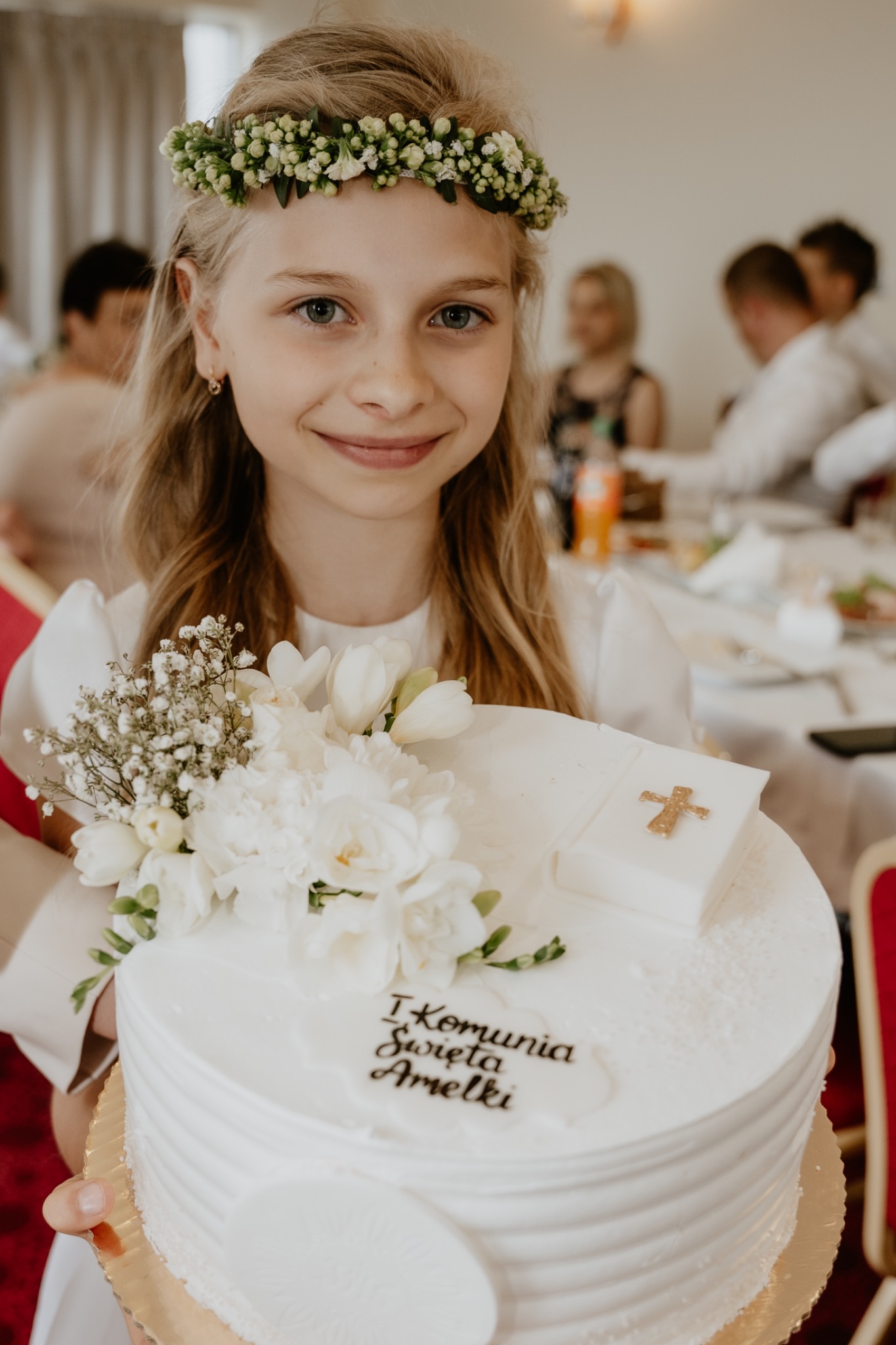 Uśmiechnięta dziewczynka w wianku z kwiatów trzyma tort komunijny z napisem 'I Komunia Święta Amelki' i dekoracją z białych kwiatów.