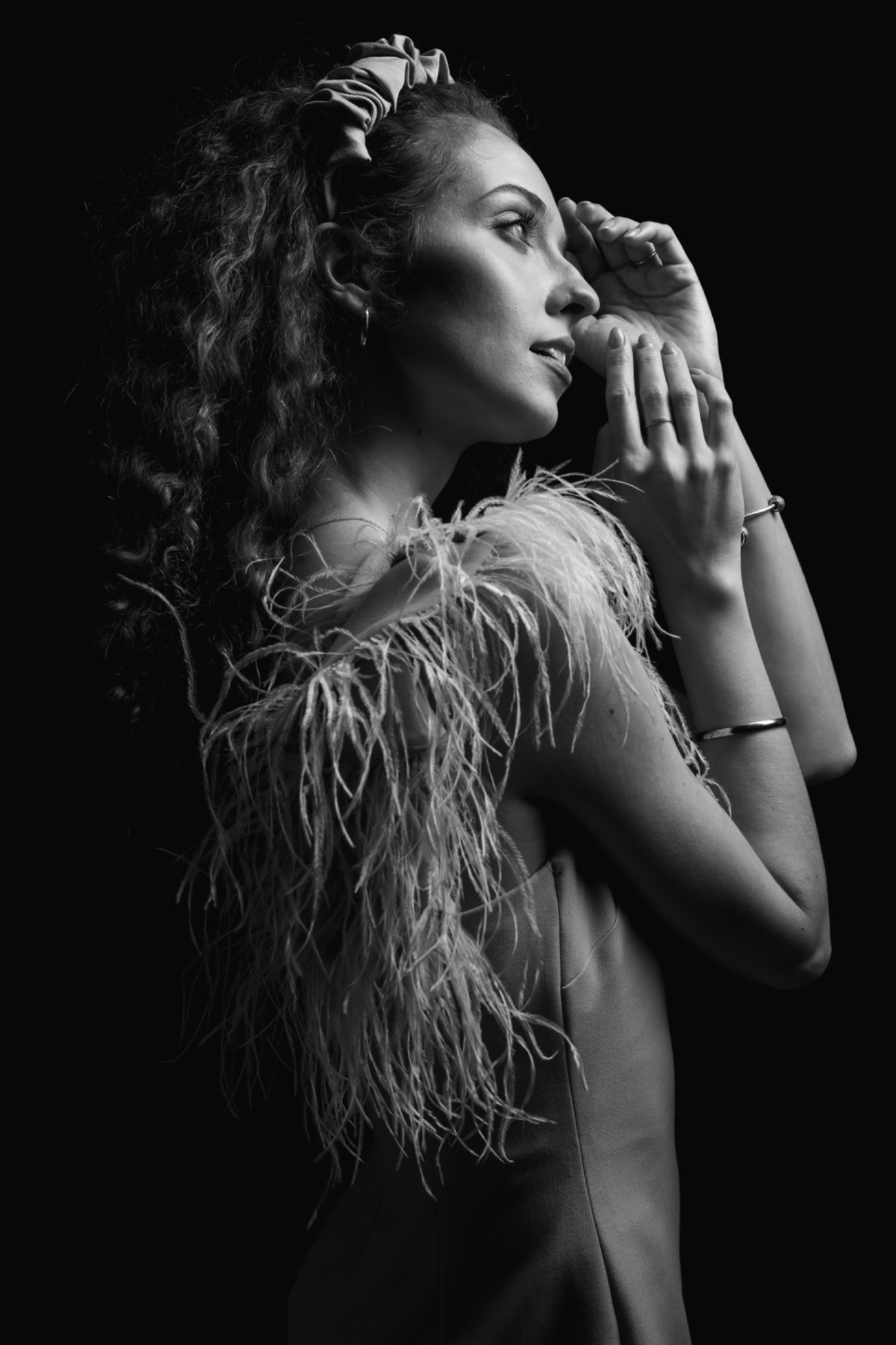 Portret kobiety w sukni z piórami, czarno-białe zdjęcie z profilu, z rękami uniesionymi do twarzy i miękkim światłem podkreślającym kręcone włosy i delikatny makijaż.