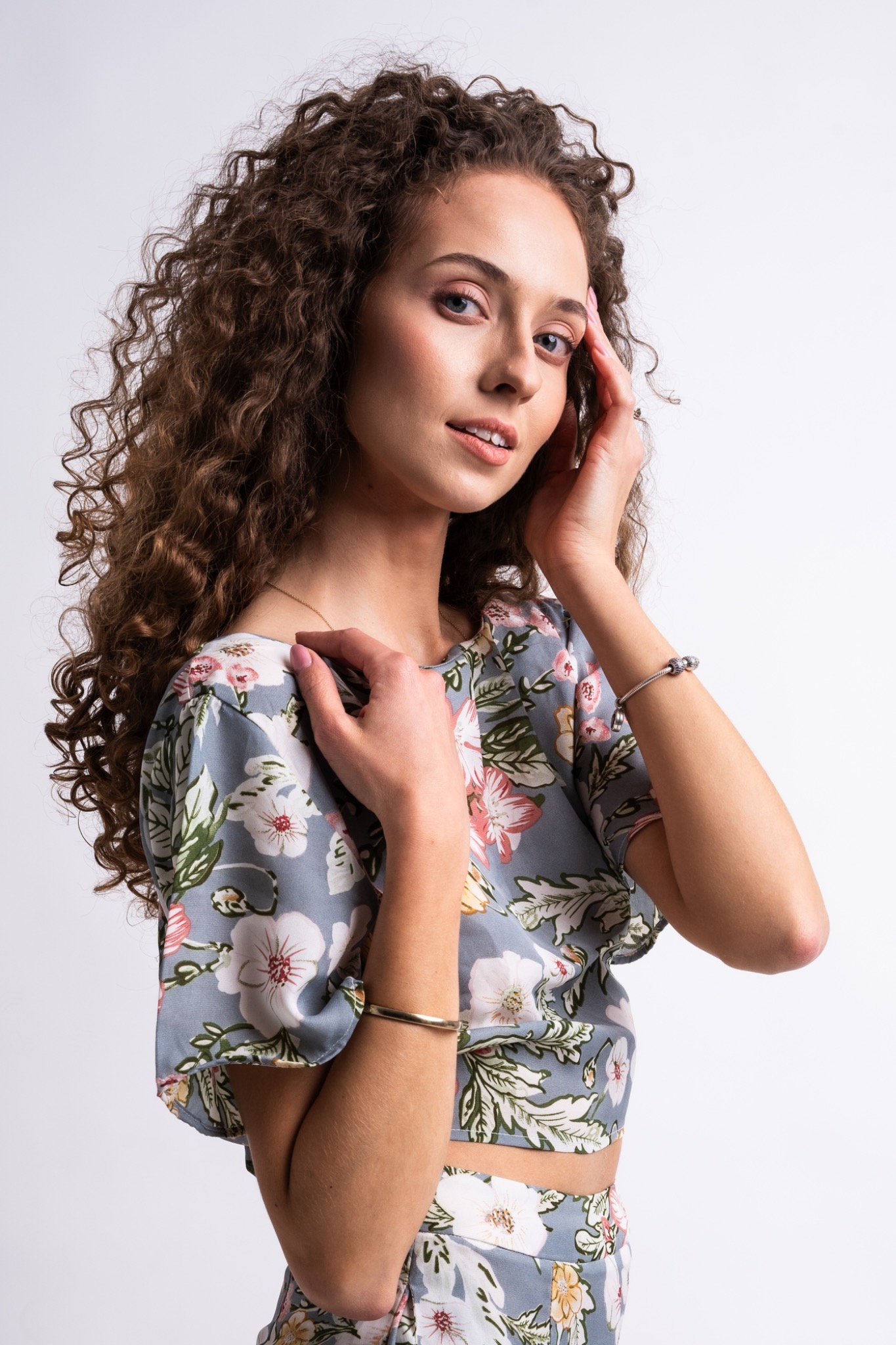 Portret młodej kobiety z kręconymi włosami w kwiecistym topie, pozującej na jasnym tle, z delikatnym uśmiechem i dłońmi dotykającymi twarzy i ramienia, eksponując biżuterię.