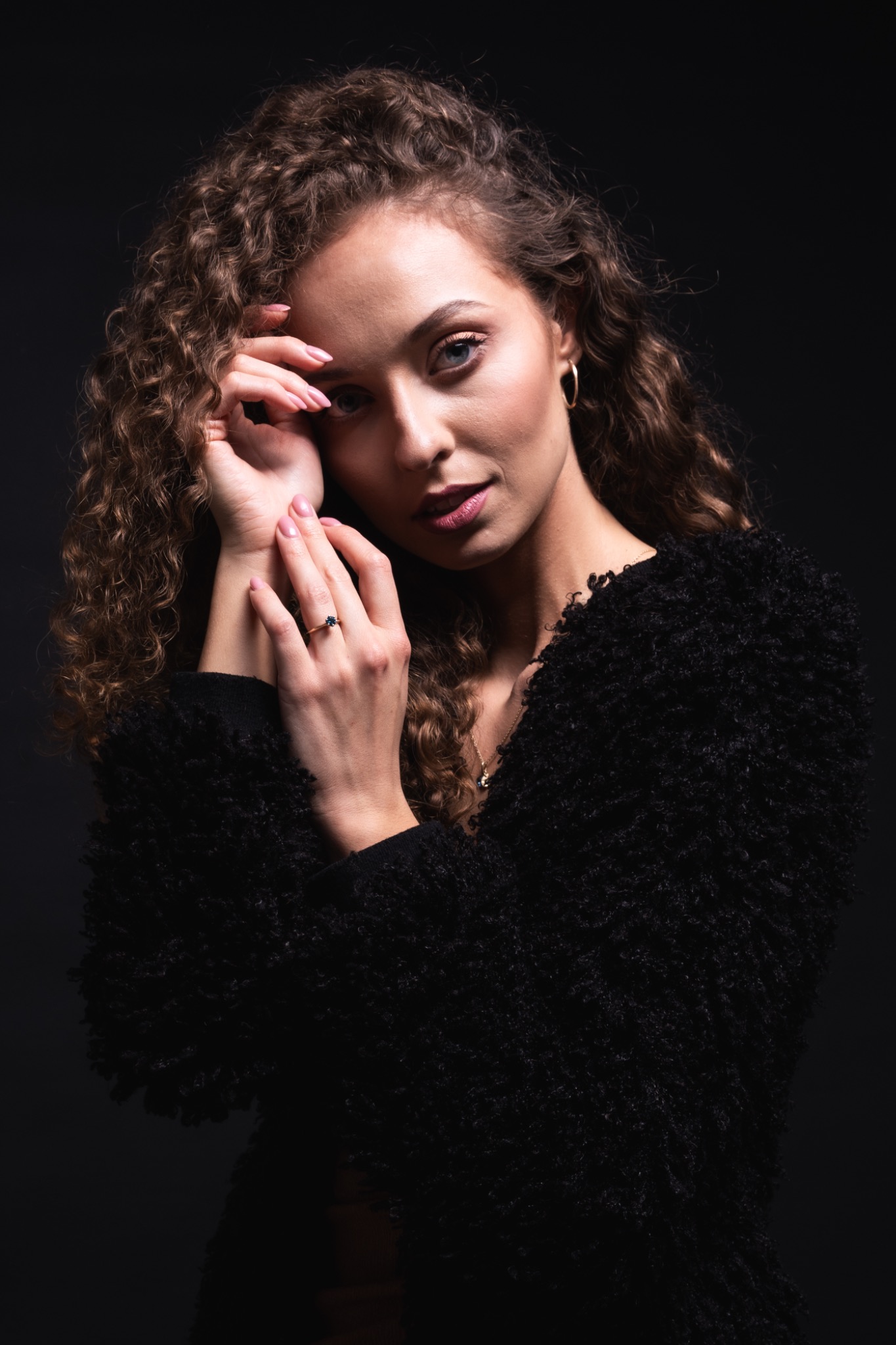 Portret kobiety z kręconymi włosami w czarnym futrzanym swetrze, delikatny makijaż, pierścionek z ciemnym oczkiem na palcu, pozuje na ciemnym tle.