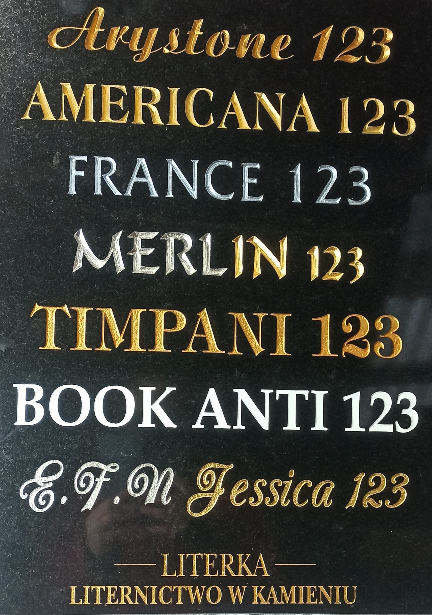 Czarna kamienna tablica z wygrawerowanymi nazwami różnych krojów pisma i numerem 123, w odcieniach złota, srebra i bieli, prezentująca liternictwo w kamieniu.