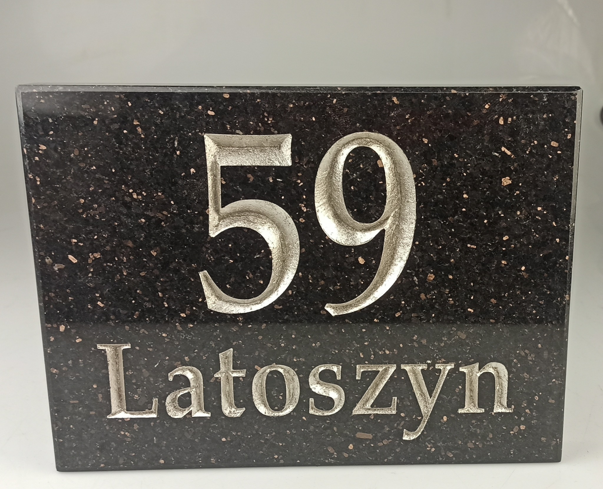 Tabliczka adresowa z czarnego, nakrapianego kamienia z wygrawerowanym srebrnym numerem 59 i napisem 'Latoszyn'.
