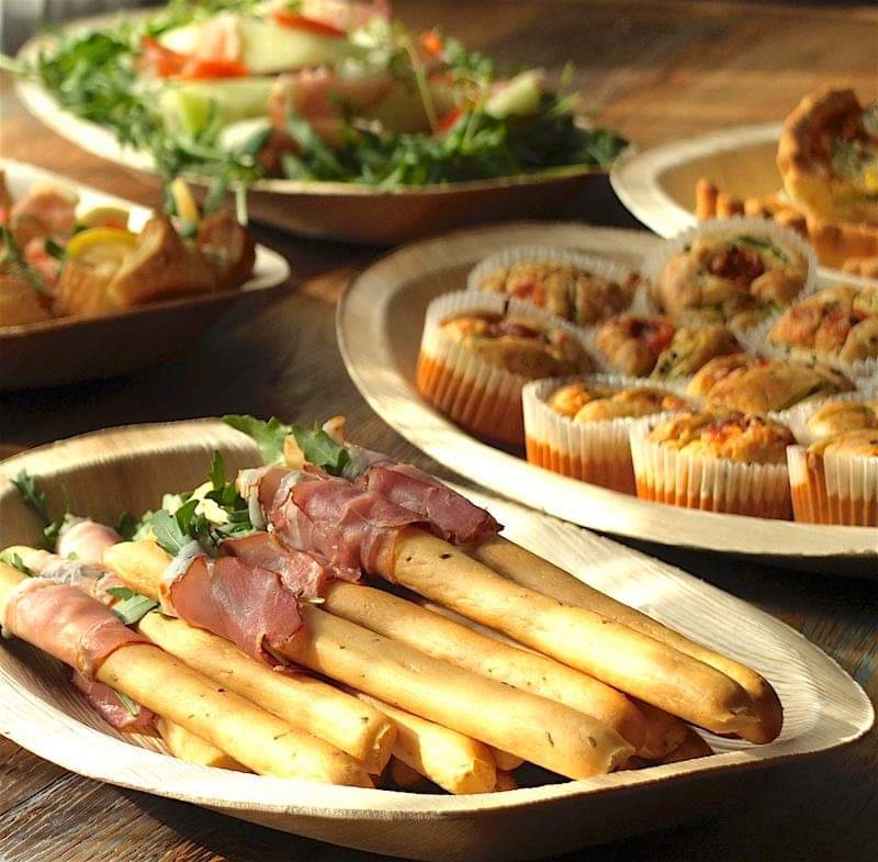 Grissini owinięte szynką parmeńską na drewnianym talerzu, obok muffiny wytrawne i sałatka z rukolą na drewnianym stole.