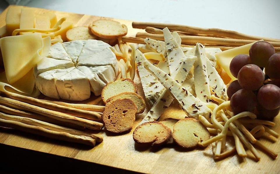 Deska serów z camembertem pokrojonym na kawałki, trójkątami sera z ziołami, grissini, gronem winogron i tostami na drewnianej desce.