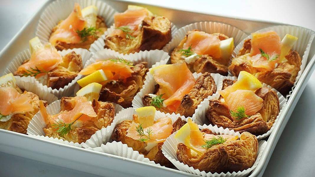 Kruche babeczki z łososiem, cytryną i koperkiem, ułożone w białej formie do transportu, widoczne detale ciasta francuskiego i dekoracji.