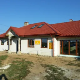 Dom w stanie surowym zamkniętym z zamontowanymi oknami o brązowych ramach i czerwonym dachem z dachówką, widoczne okna dachowe, kolumny wejściowe owinięte folią ochronną, teren wokół w trakcie...