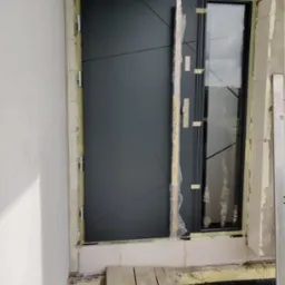 Montaż nowoczesnych drzwi wejściowych z bocznym przeszkleniem, widoczne pianka montażowa i deski zabezpieczające podłoże, budowa w trakcie wykończenia.