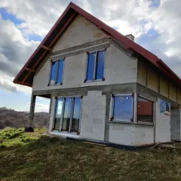 Surowy budynek jednorodzinny w trakcie montażu okien z brązowymi ramami, widoczne bloczki betonowe i czerwona dachówka, krajobraz pagórkowaty w tle.