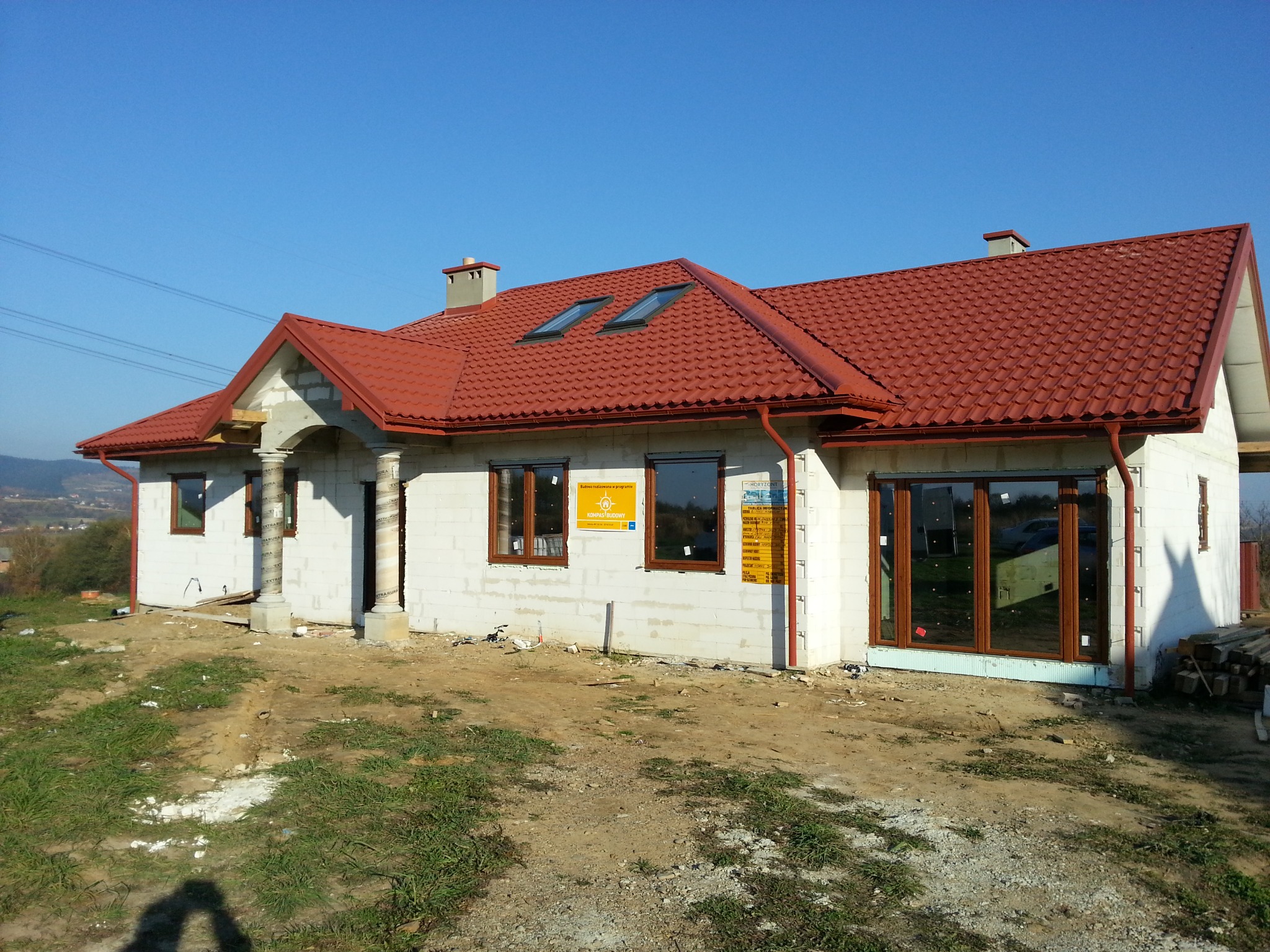 Dom w stanie surowym zamkniętym z zamontowanymi oknami o brązowych ramach i czerwonym dachem z dachówką, widoczne okna dachowe, kolumny wejściowe owinięte folią ochronną, teren wokół w trakcie...