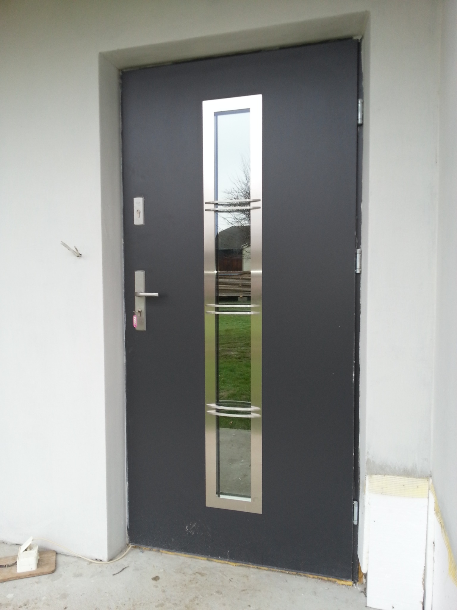 Nowoczesne, antracytowe drzwi wejściowe z długim, pionowym przeszkleniem ze stali nierdzewnej, widoczne z zewnątrz, z fragmentem ocieplenia ściany obok.