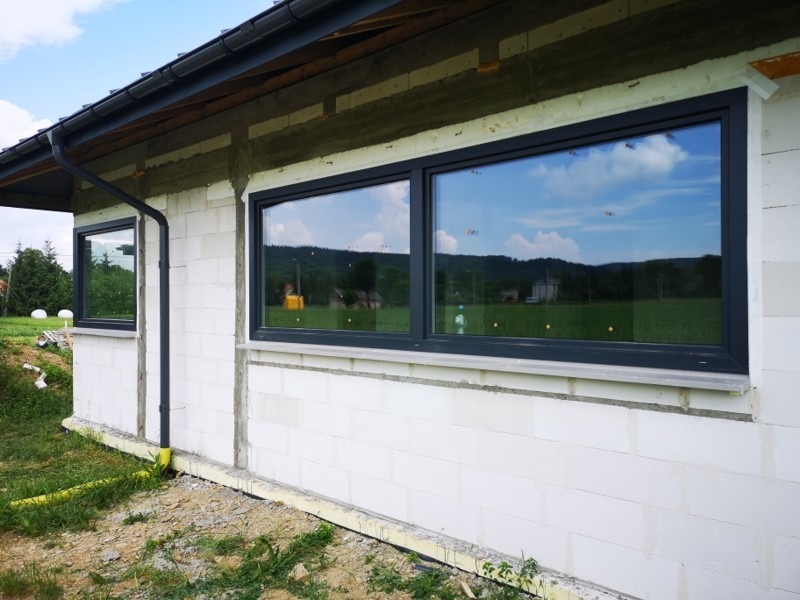 Surowy stan budynku z białego bloczka z zamontowanymi grafitowymi oknami z widokiem na krajobraz i odbiciem nieba w szybach. Widoczny fragment rynny i niedokończone ocieplenie.